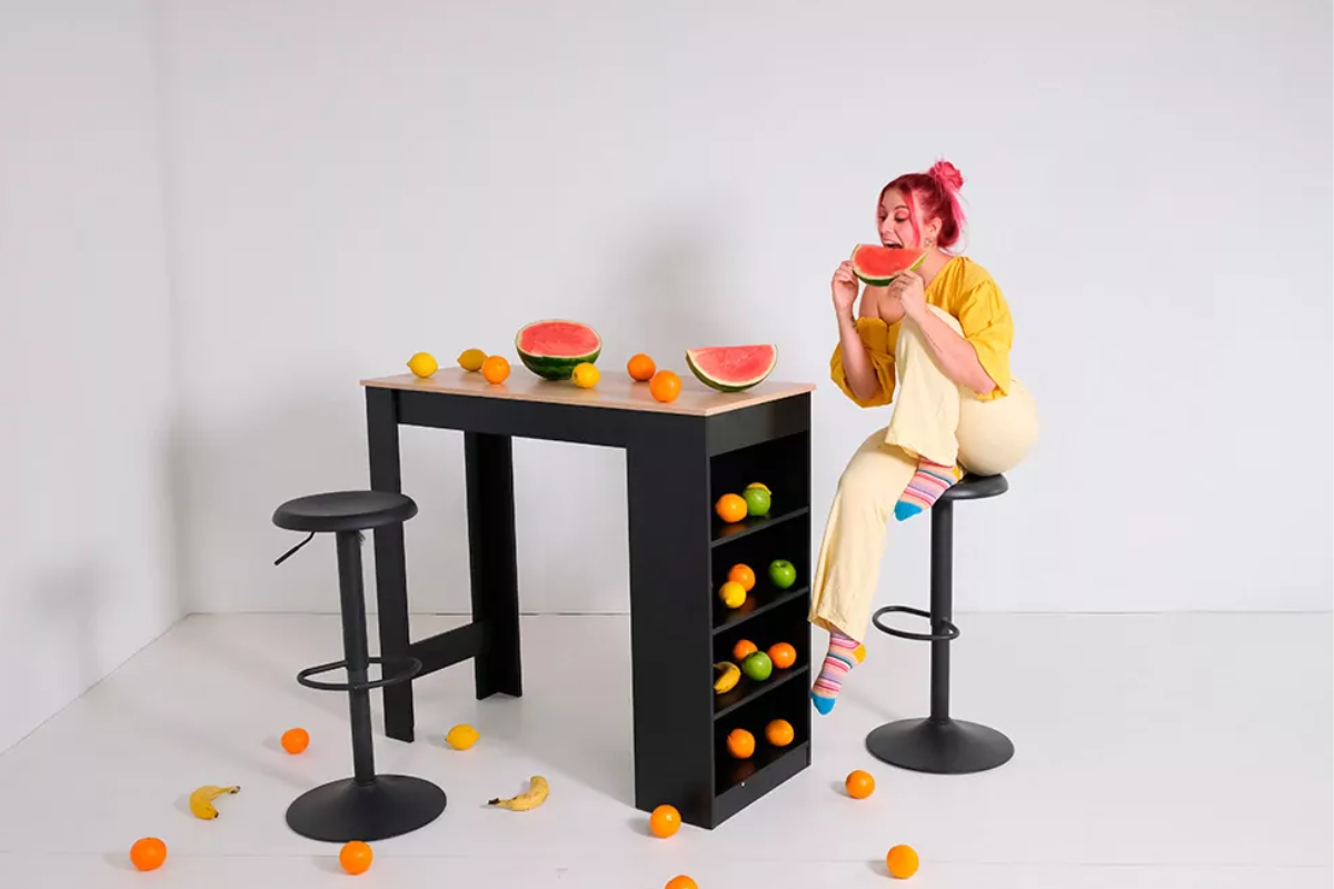 Mesa alta negra con estantes y taburetes, mujer sentada comiendo fruta en la mesa