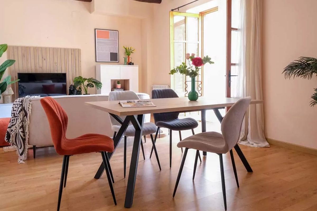 Mesa de comedor rectangular fija, con tono amaderado. Decorada con sillas de varios colores y un jarrón con flores encima