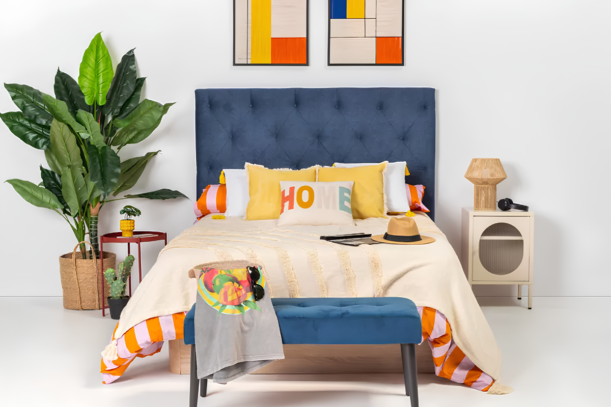 Dormitorio decorado con cama de matrimonio con cabecero tapizado, dos cuadros encima y a los lados una mesita de noche y una planta artificial. Delante de la cama un banco tapizado de color azul.