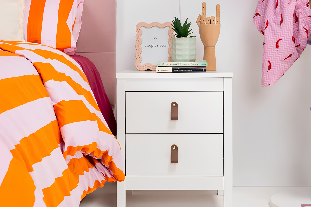 mesita de noche blanca de dos cajones, colocada al lado de una cama de matromonio, con objetos decorativos encima como marcos de fotos